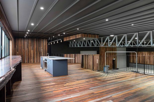 A spacious industrial-style interior with a large kitchen island, extensive timber flooring and vertical timber wall cladding. The ceiling features exposed grey metal beams and linear ribbed detailing, with track and recessed lighting. A prominent steel truss spans the upper level, while black metal balustrades frame an open stair or mezzanine edge. Large windows line one side, and a substantial live-edge timber counter is visible at the left.