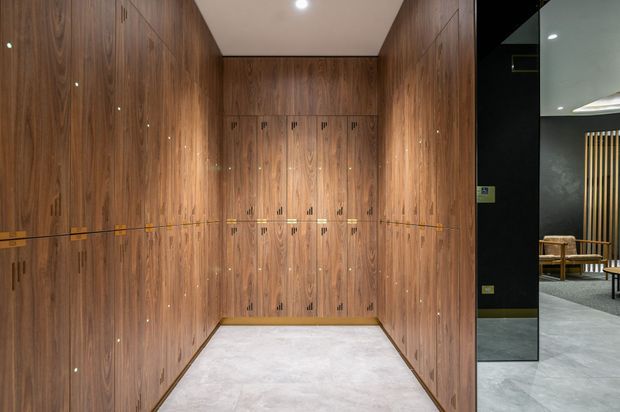 A compact, highly finished timber-lined room or storage area with floor-to-ceiling dark wood cabinetry on three sides, integrated vertical handles, and a pale stone or concrete-look floor. A glazed opening at right reveals part of an adjacent contemporary lounge or showroom with dark walls, light grey flooring, and timber furniture.