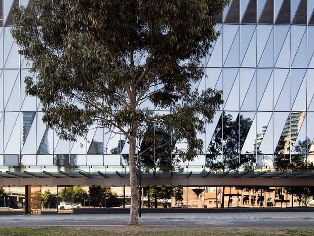 Street-facing exterior view of a contemporary multi-storey commercial building with a highly reflective geometric glazed facade, dark horizontal canopy, and transparent ground-level frontage. A mature street tree partially obscures the building, with reflected trees and nearby structures visible in the glazing.