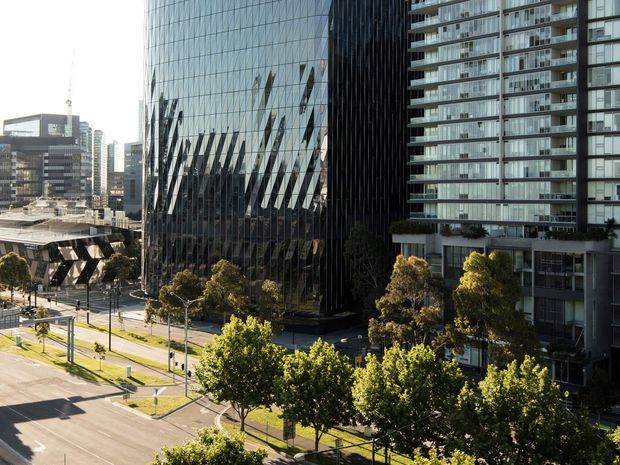 Urban cityscape viewed from an elevated position, showing a prominent contemporary glass office tower with reflective facades, adjacent high-rise residential buildings, landscaped street trees, broad roads, and surrounding commercial development.