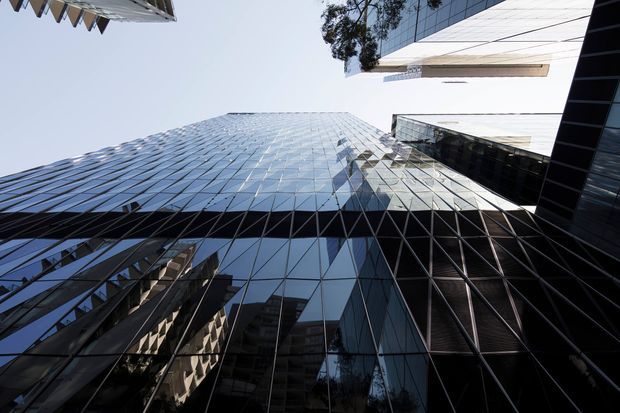 Upward-looking architectural photograph of a dense urban cluster of tall contemporary office towers. The frame is dominated by reflective blue-grey glass curtain walls, dark metal mullions, sharp geometric facades, and projecting white structural or cladding elements, creating a dramatic canyon-like perspective against the sky.