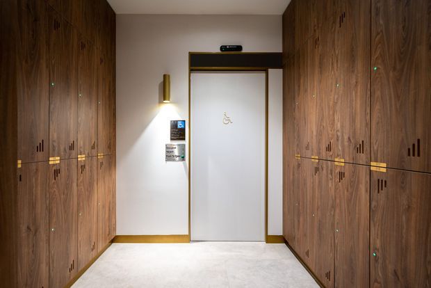 A compact, refined corridor or lift lobby lined on both sides with extensive dark timber veneer cabinetry or wall panelling. The central accessible lift door has a crisp white finish with slim brass-gold trim, beside a wall-mounted cylindrical light and accessibility signage. The space has a pale smooth floor, warm recessed lighting, and subtle black ventilation or handle details integrated into the timber panels.