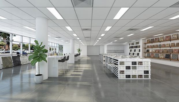 Bright, spacious contemporary commercial showroom or design materials retail space with a polished grey floor, white cylindrical columns, extensive ceiling grid lighting, floor-to-ceiling windows, central white display islands, shelving with sample boards, and a small café-style consultation counter. Minimal white and grey fit-out with indoor plants and abundant daylight.