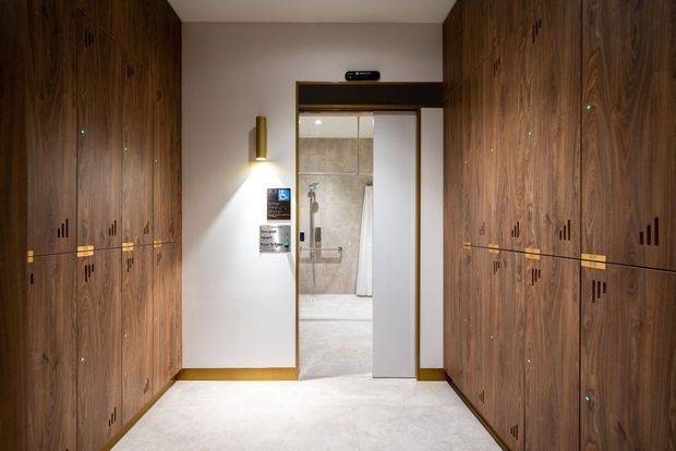 A refined interior hallway or entry corridor lined on both sides with extensive dark timber built-in cabinetry. The cabinetry features integrated brass-toned horizontal details, black ventilation or handle motifs, and small green indicator lights. A wall-mounted brass light fixture illuminates the pale plaster wall, while a doorway with brass trim opens to a tiled bathroom or washroom beyond. The floor is a light polished stone or concrete-like finish, creating a warm, elegant, highly organised space.