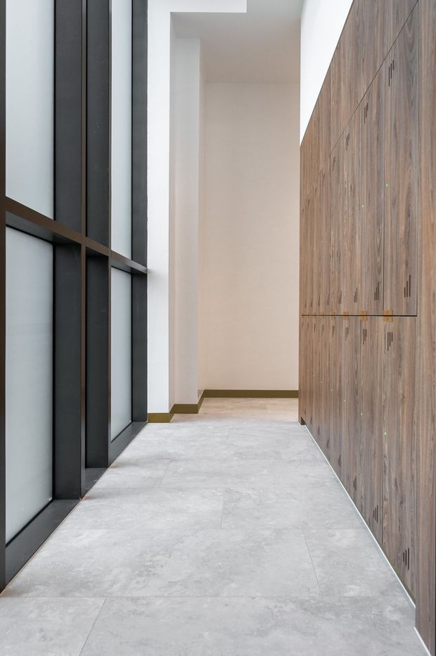 A narrow, minimalist interior corridor or entry passage with full-height black-framed glazing on the left, a continuous wall of warm wood built-in cabinetry on the right, pale stone-look flooring, and white walls. The clean linear composition emphasizes the tall glazing, concealed storage, and restrained material palette.