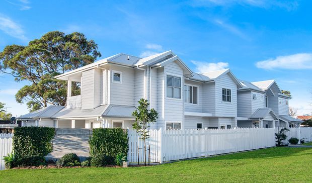 Exterior view of a large white multi-storey residential building with repeated gabled roof sections, horizontal weatherboard cladding, white-framed windows, covered balconies, and a white picket fence bordering a manicured lawn. The architecture has a bright coastal/suburban character.