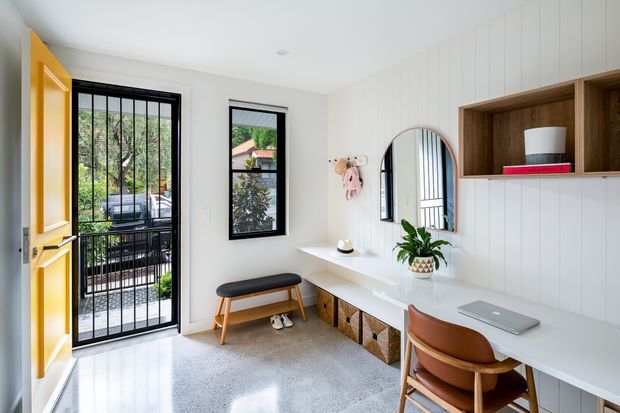 Bright, compact entryway with a built-in home office and storage. The space features a white vertical-panel wall, terrazzo flooring, a long white built-in desk and bench, warm timber cabinetry, black-framed glazing and security door, and a rounded wall mirror. Natural light creates a clean, contemporary and inviting atmosphere.