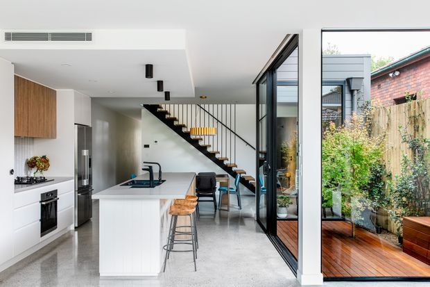 Bright open-plan contemporary kitchen and dining area with a white island, integrated appliances, timber cabinetry, and bar stools. A black metal stair with timber treads leads upward beside full-height glazing, which opens views to a timber deck and planted courtyard. The space has polished concrete flooring and a clean, modern material palette.