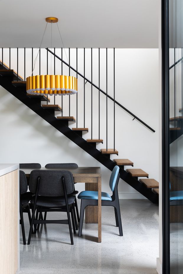 A contemporary dining area beneath a dramatic open-riser staircase. The staircase has a black steel stringer and balustrade with slim vertical metal rods, timber treads, and a minimalist handrail. A striking gold cylindrical pendant light hangs above a dark timber dining table with charcoal upholstered chairs and a blue accent chair. Polished grey concrete flooring, white walls, and black-framed glazing complete the modern interior.