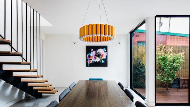Contemporary dining area with a long dark timber dining table, upholstered dining chairs, sculptural orange pendant light, white walls, and a dramatic open-riser staircase with black metal structure. Full-height glazing opens to a timber deck and enclosed garden, with colourful artwork on the wall.