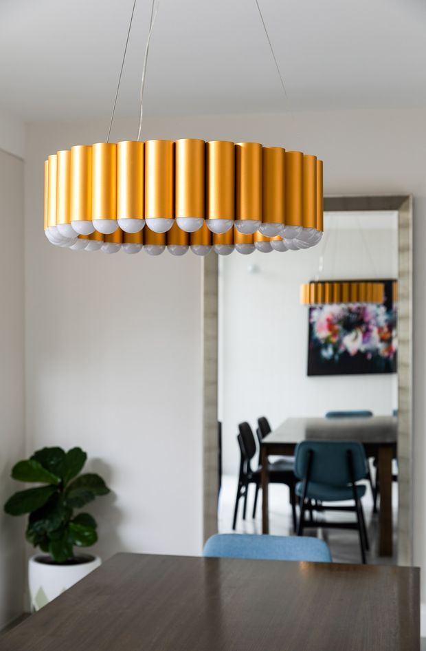 Interior view dominated by a sculptural gold pendant light above a dark timber dining table. A large framed mirror reflects a dining area with dark chairs and colourful wall artwork, while a potted plant sits against a light neutral wall.