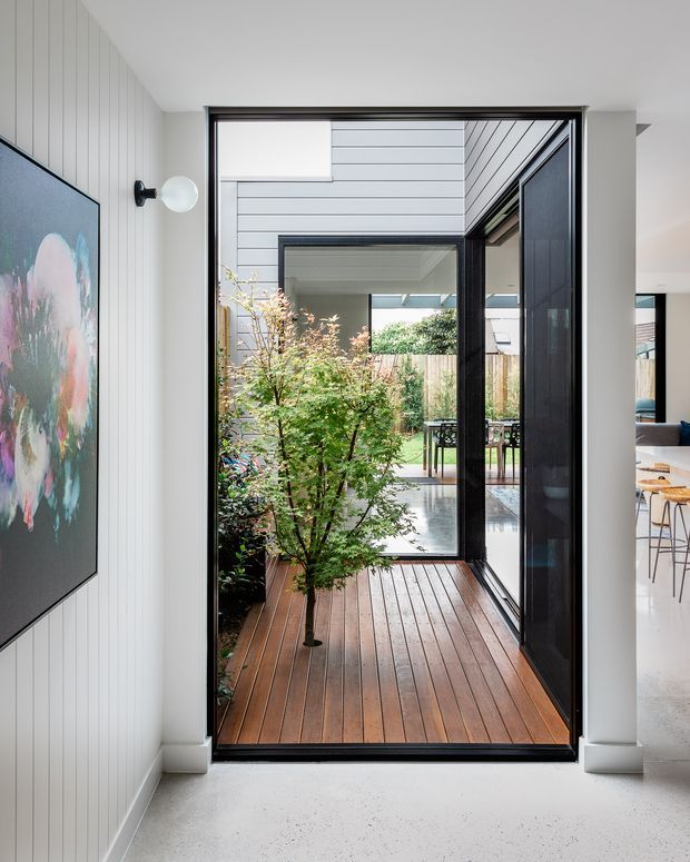 Bright contemporary interior looking through a full-height black-framed glazed doorway onto a timber deck and landscaped courtyard. The entry area has white vertical wall panelling, a polished light floor, a wall-mounted globe light, and a large colourful artwork. A dining area is partially visible to the right, while the courtyard includes greenery, fencing, and a covered outdoor seating area.