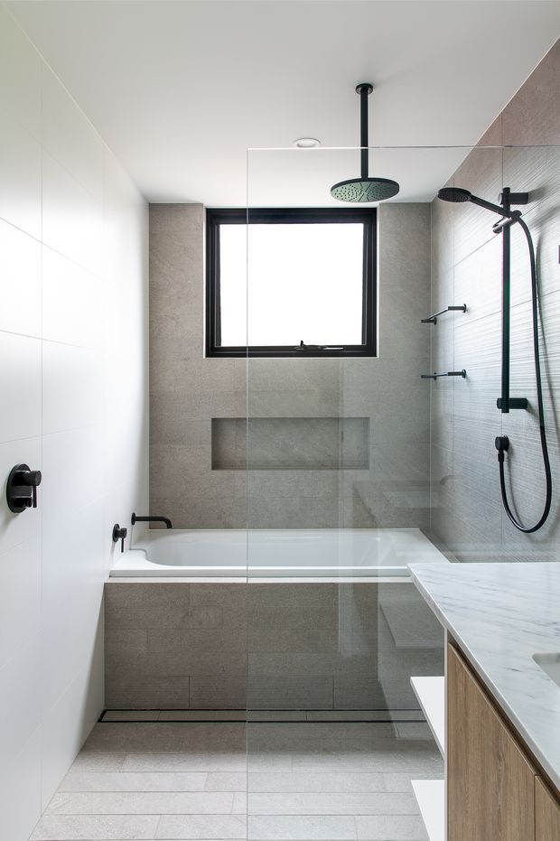 A bright contemporary bathroom with a bathtub and overhead shower enclosed behind a frameless glass screen. Large pale grey stone-look tiles cover the walls and floor, with textured wall tiles on the shower side. Matte black tapware, shower fittings and window framing contrast with the light finishes. A timber-veneer vanity with a pale stone benchtop is visible in the foreground.