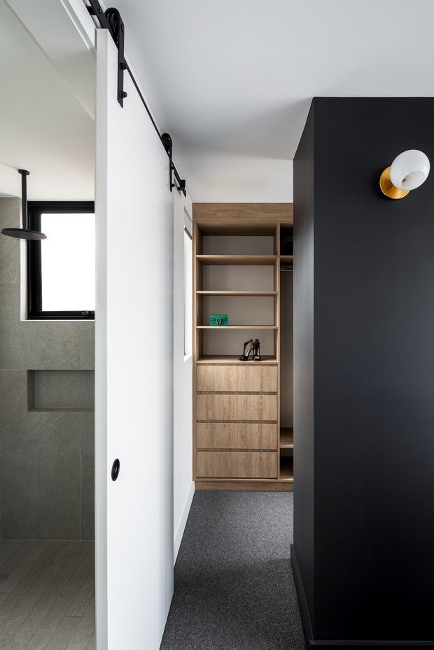 A compact modern dressing and circulation area viewed from a bathroom. A white sliding barn-style door with black hardware opens toward a built-in timber wardrobe with open shelving and drawers. Dark charcoal cabinetry or wall joinery forms a strong foreground feature, with a black-and-gold wall sconce. The adjacent bathroom has grey stone-look wall and floor tiles, a black-framed window, and a recessed shower niche.