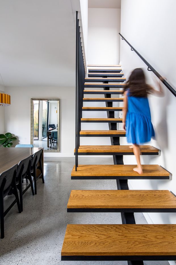 Bright contemporary open-plan interior featuring a floating staircase with thick warm timber treads, black central steel support, and slim black balustrade. A dining area with a dark table and chairs is visible beside the stair, with white walls, polished grey concrete flooring, and a large wall mirror.