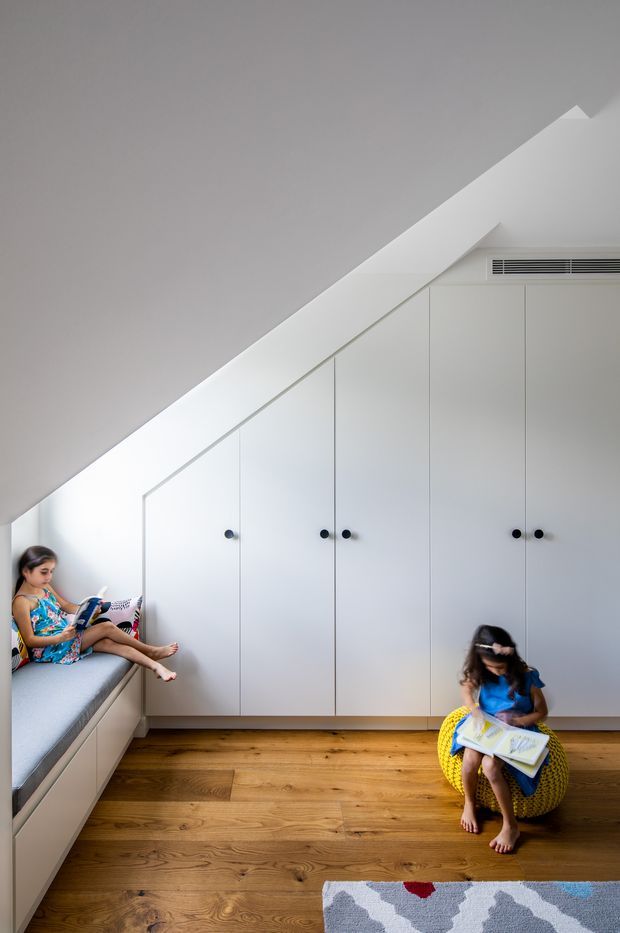 Bright, minimalist children's room beneath a sharply sloping ceiling, with extensive custom white built-in cabinetry following the roofline, integrated window seating, warm timber flooring, and two children reading. A patterned rug and yellow woven chair add playful colour.