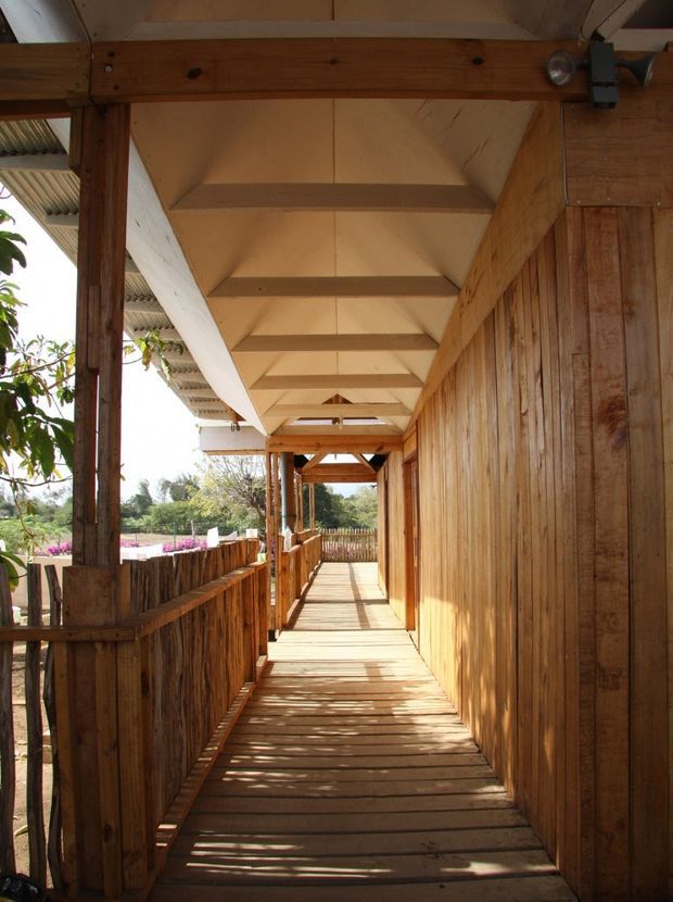A narrow covered exterior walkway or veranda constructed predominantly from timber. The image shows a timber plank floor, vertical timber-clad wall on the right, timber balustrades and posts on the left, and exposed timber roof framing overhead. Strong sunlight casts linear shadows across the deck, with garden vegetation visible beyond the railing.
