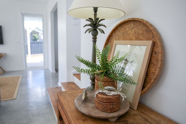 A styled entryway or hallway vignette with a rustic timber console table, woven baskets, a large round woven wall piece, framed artwork, greenery, and a decorative tropical-style table lamp. A bright corridor and doorway extend into the background, with pale walls and a polished light floor.