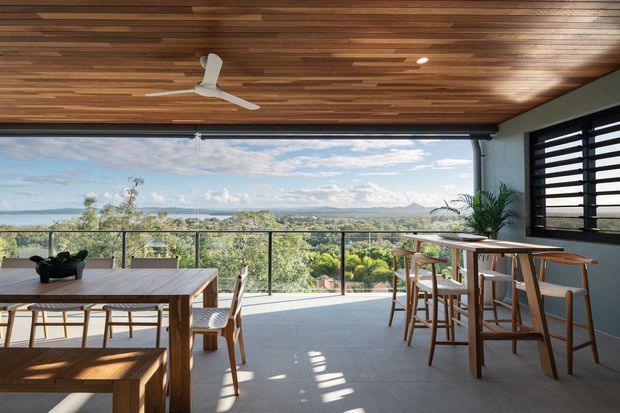 A bright, open-plan dining and entertaining area with a large timber dining table and bench, a timber bar-height counter with stools, full-width retractable glazing, dark louvred windows, and a striking timber-lined ceiling. The room opens onto a balcony with expansive views of tropical vegetation, water, and distant hills. A white ceiling fan and recessed lighting complete the relaxed contemporary setting.