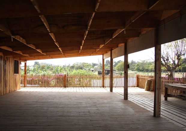 A spacious covered outdoor timber deck or veranda with a large exposed timber roof structure, timber posts, plank flooring, and rustic vertical slat balustrades. The open-sided space overlooks a leafy residential landscape and includes a simple timber dining table at the right.