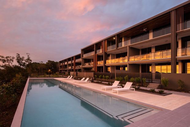 A modern multi-storey residential building viewed at dusk from a long swimming pool terrace. The building has a strong horizontal form, dark timber-toned soffits and facade elements, extensive glazing, recessed balconies, and vertical screening details. The pool and pale paved deck occupy the foreground, with landscaped planting along the building.