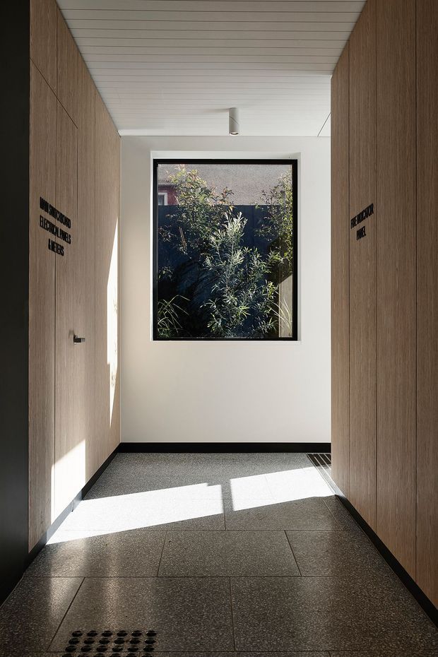 A narrow, minimalist interior corridor or entry passage with full-height warm timber wall panelling, a white slatted ceiling, pale plaster end wall, and a black-framed window overlooking planted greenery. Large dark grey floor tiles, black skirting, recessed drainage detail, and strong sunlight create a clean, refined institutional or commercial entrance.