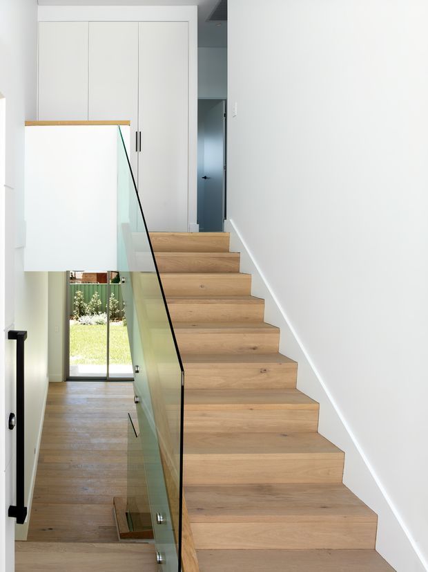 Bright minimalist stairway with a straight run of pale timber stairs, clear glass balustrade with slim black edging, white walls, and integrated white cabinetry at the upper landing. A lower hallway and glazed door to a garden are visible beyond the balustrade.