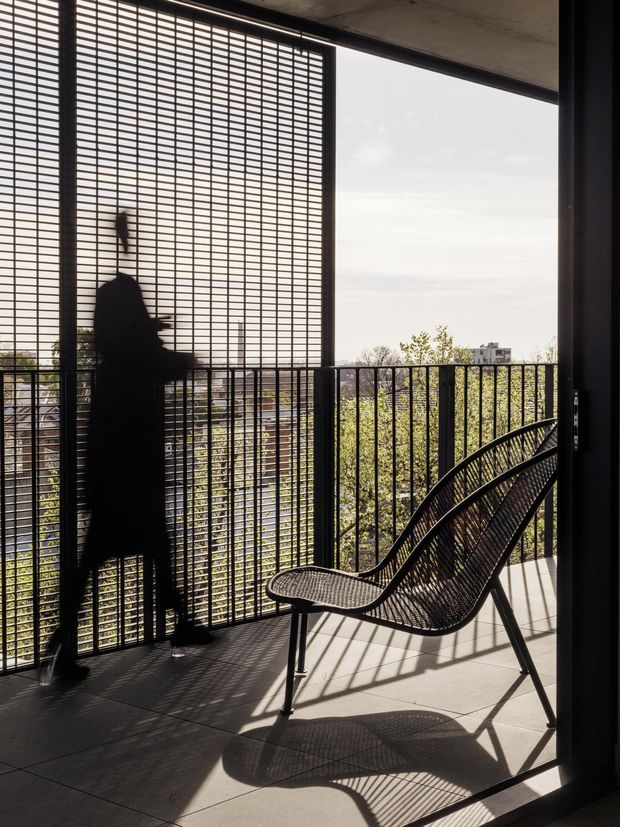 A sunlit contemporary balcony or terrace viewed from indoors, enclosed by black metal railings and a large gridded metal privacy screen. A black woven lounge chair sits on large pale floor tiles, casting strong geometric shadows. A silhouetted person stands beside the screen, with trees and urban buildings visible beyond the balcony.