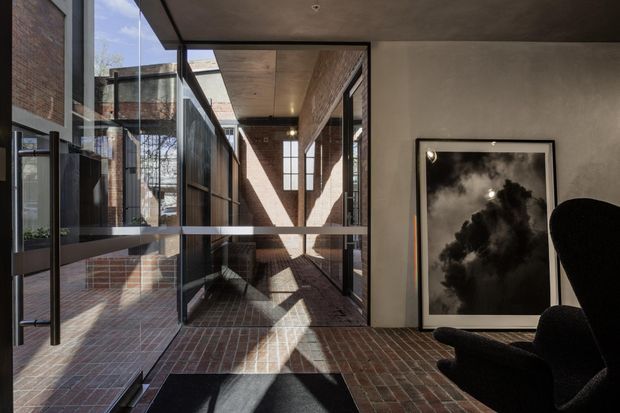 A dramatic contemporary interior with a glazed corridor or entry space framed by exposed red brick walls, dark metal window and door frames, and a dark ceiling. Strong sunlight casts geometric shadows across the brick floor and walls. A black lounge chair and monochrome artwork sit to the right, while extensive glazing reflects the adjoining brick-lined spaces.