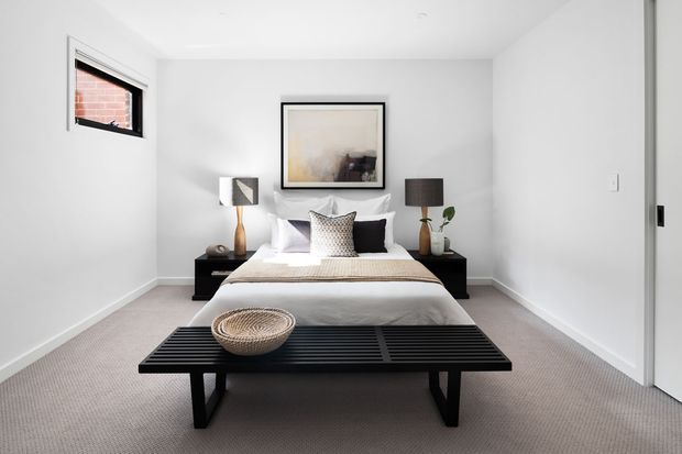 A bright, minimalist bedroom with a neatly made bed centered against a white wall, symmetrical bedside tables and lamps, framed abstract artwork, and a black slatted bench at the foot of the bed. The room has soft neutral furnishings, light carpet, white walls, and a small high-set window.