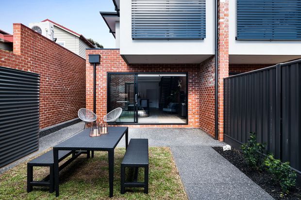 Contemporary urban courtyard at the rear of a brick townhouse, featuring a compact gravel-paved outdoor dining area with a black picnic-style table and benches, black metal fencing, exposed red brick walls, and large glazed doors opening into the interior. Upper-level windows have dark horizontal external sunshades.