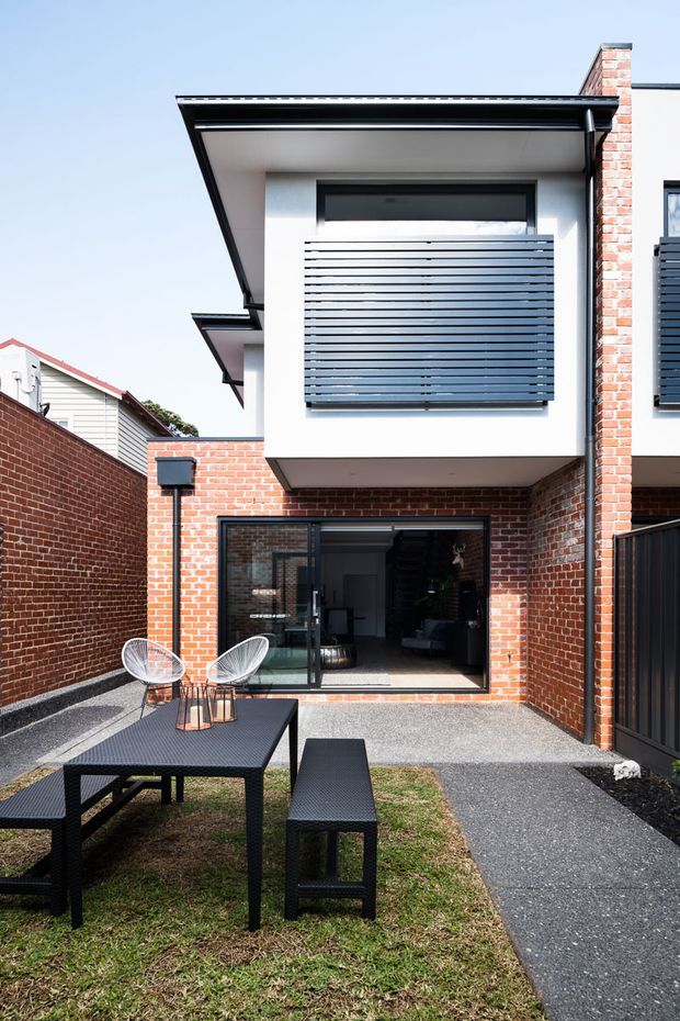 A compact modern multi-storey townhouse courtyard viewed from the outdoor area. The facade combines exposed red brick walls with white rendered upper walls, black metal roof trim, dark horizontal privacy screens, and large glazed openings. A paved path leads through a small grass courtyard furnished with a black outdoor dining table, bench seating, and wire-frame chairs. A dark interior stairway and living area are visible through the ground-floor glazing.