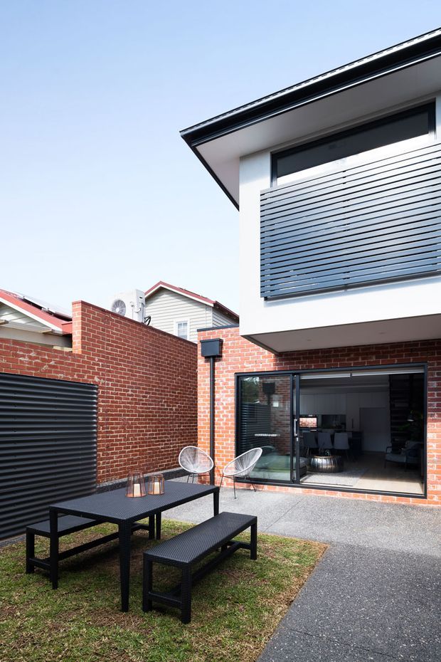 A contemporary residential courtyard viewed from outdoors, featuring a red brick wall, black metal outdoor dining table and benches, a paved path, and a modern two-storey extension. The building has white rendered walls, dark horizontal privacy screens, black-framed glazing, and a projecting upper level. A living area is visible through the open sliding doors.