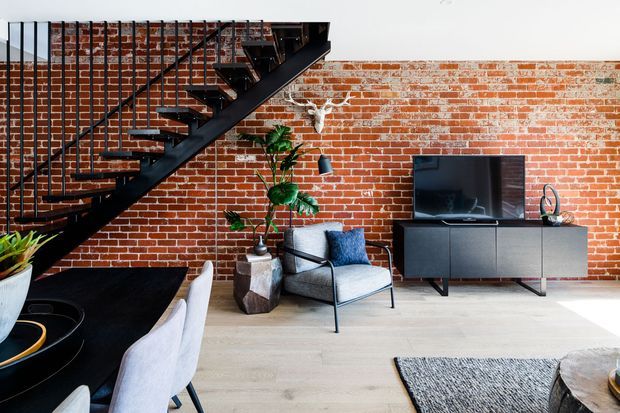 Industrial-style living room with an exposed red brick feature wall, black open-riser staircase and metal balustrade, pale timber flooring, and a compact media console with television. A contemporary lounge chair, dining area, indoor plant, and decorative objects complete the space.
