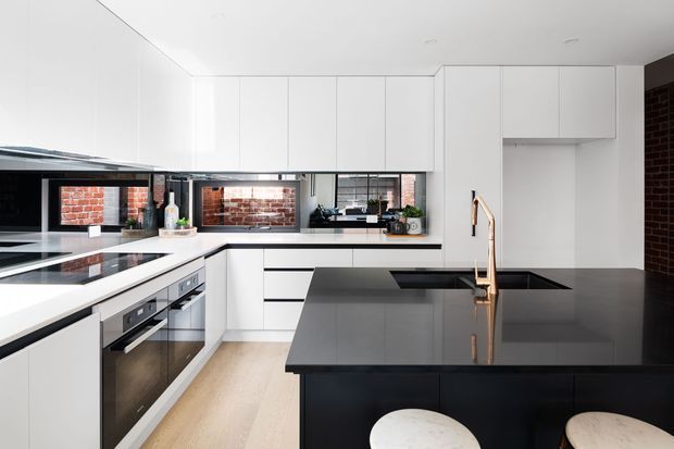 Contemporary kitchen with white flat-panel cabinetry, a large dark stone island with integrated sink and bronze tapware, light timber flooring, black appliances, and exposed red brick visible through the window openings and at the side wall.