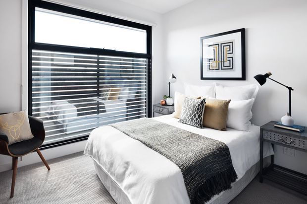 Bright, contemporary bedroom with a neatly made bed, layered white bedding, textured grey and black throw, patterned accent cushions, and matching dark bedside tables. A large black-framed window with horizontal blinds provides natural light, while geometric wall art and a sculptural armchair add restrained decorative detail.