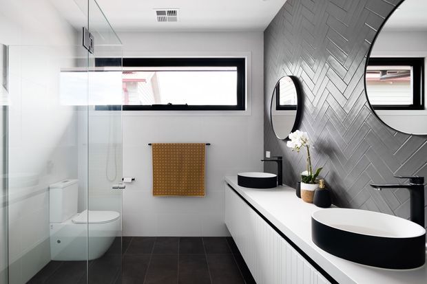 Contemporary bathroom with a walk-in glass shower, wall-hung toilet, dark tiled floor, floating double vanity, vessel basins, and round mirrors. A grey herringbone feature wall contrasts with white cabinetry and fixtures, while a narrow horizontal window provides natural light.