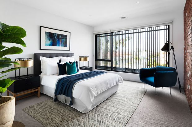 A bright, contemporary bedroom with a neatly made bed, white bedding, dark navy throws and teal accent cushions. The room features a large black-framed floor-to-ceiling window with vertical blinds, an exposed brick feature wall, dark bedside furniture, a blue armchair, a black floor lamp, a textured area rug, and a large potted plant.