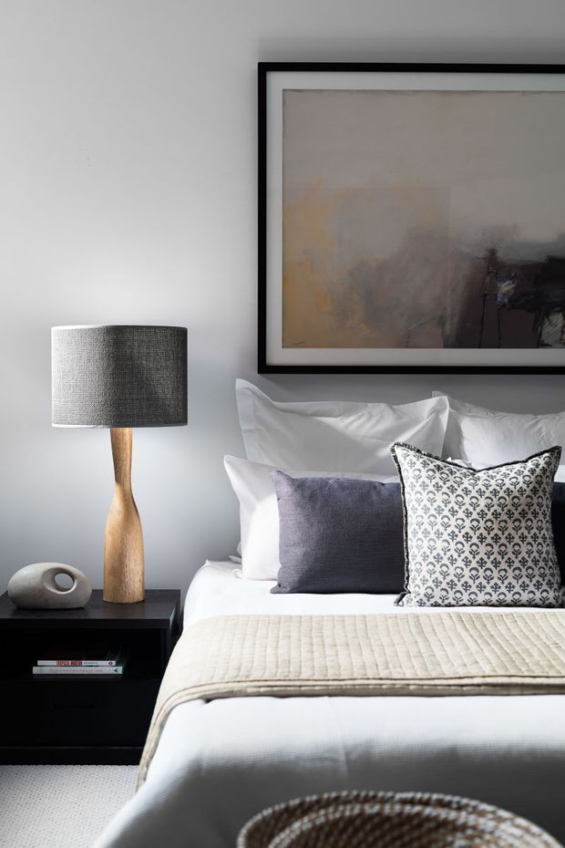 A refined bedroom vignette with a neatly made bed, layered white and patterned pillows, a pale textured throw, and a dark bedside table. A sculptural wooden lamp with a grey fabric shade sits beside the bed, while abstract artwork in a black frame hangs on the soft grey wall. The room has a calm, contemporary and subtly luxe appearance.