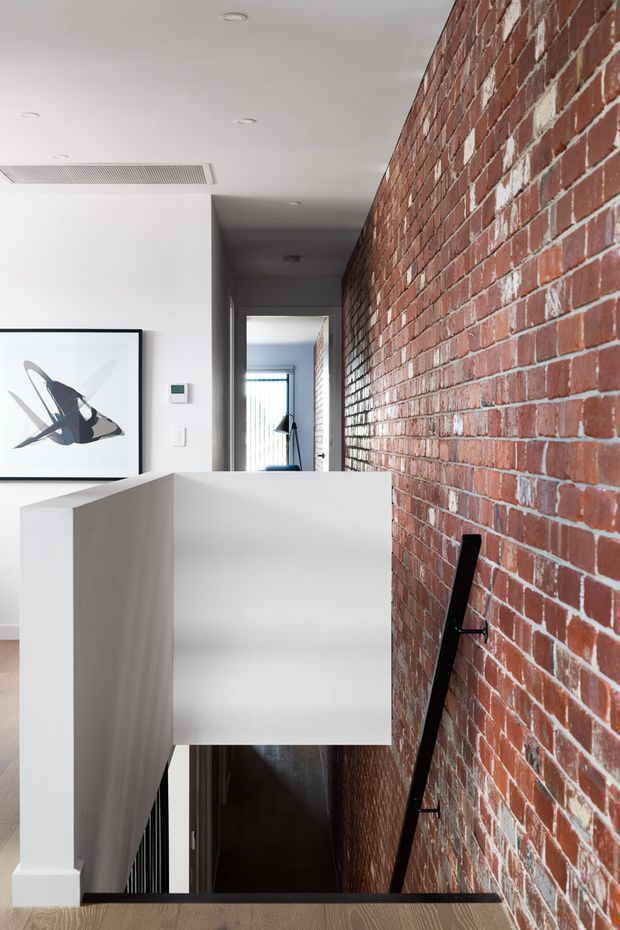 Interior view of a narrow stairway and hallway with a strong exposed red brick feature wall, white plastered walls and ceiling, and a minimalist white balustrade. A black handrail descends along the brick wall toward the lower level. Recessed ceiling lights, a framed abstract artwork, and a glimpse of a bright room beyond complete the contemporary residential interior.