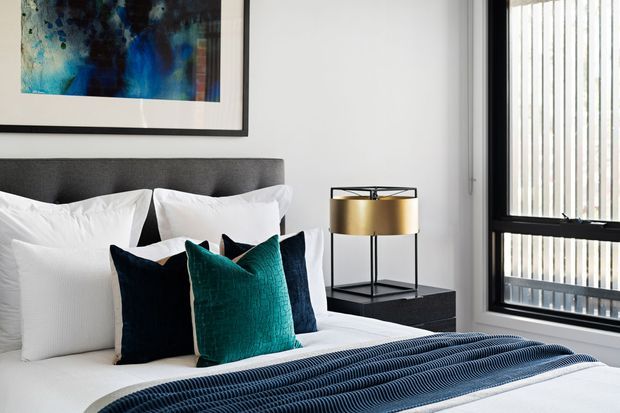 A refined contemporary bedroom with a charcoal upholstered headboard, crisp white bedding, layered navy and teal cushions, and a textured dark blue throw. A framed abstract artwork hangs above the bed, beside a black bedside table with a brass drum lamp. Large black-framed windows with vertical blinds provide strong natural light.