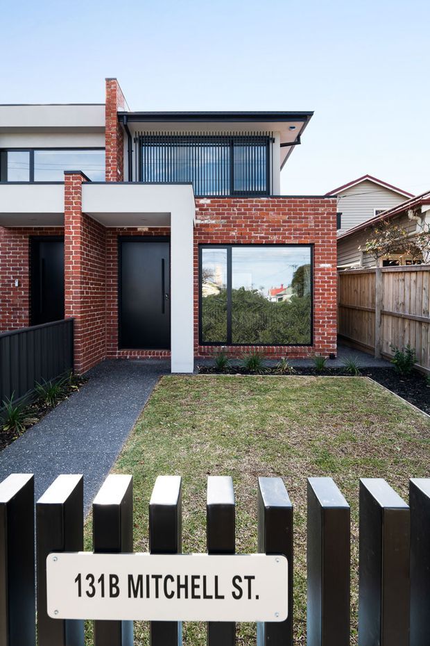 Front exterior view of a contemporary two-storey townhouse with a red brick facade, white rendered projections, dark metal-framed windows and doors, and a small landscaped front yard. A black vertical-slat fence and paved path lead to the entry, with a visible address plaque reading 131B Mitchell St. The house has a low-pitched roof and an upper-level screened balcony or window zone.