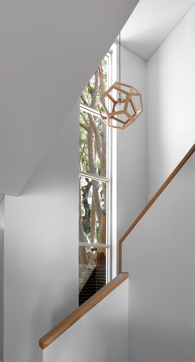 A bright, minimalist stairway interior with crisp white walls and angular ceiling planes. A tall, narrow window brings in views of mature trees, while warm timber handrails trace the staircase. A geometric timber pendant light hangs in the stairwell, adding a sculptural natural accent.
