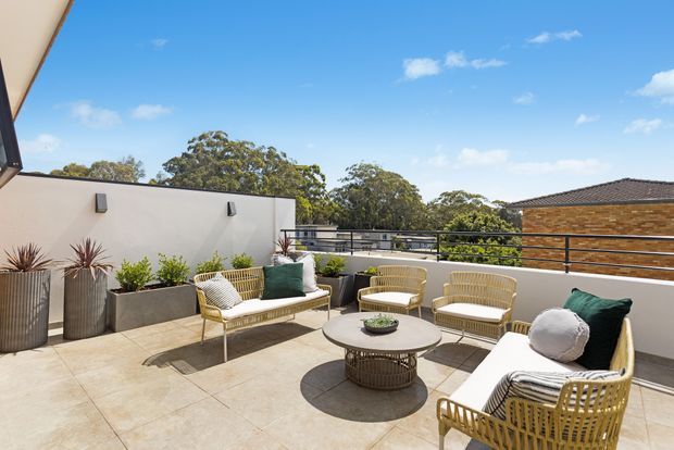 A bright rooftop terrace with expansive tiled flooring, cream rendered parapet walls, black metal railings, and a relaxed outdoor seating arrangement. Woven rattan-style chairs and a sofa surround a round coffee table, with raised planters and potted plants along the edges. Trees, neighbouring rooftops, and a clear blue sky are visible beyond the terrace.