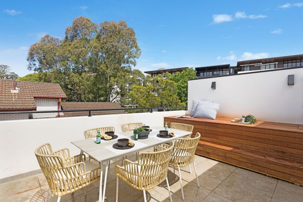 Sunlit rooftop terrace with a paved dining area, light outdoor dining table and woven chairs, and a built-in timber bench against a white rendered privacy wall. Nearby rooftops, mature trees, and modern buildings are visible beyond the terrace.