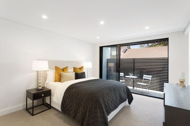 Bright contemporary bedroom with a large bed dressed in white linen and a dark charcoal quilt, mustard and charcoal accent cushions, bedside tables with lamps, and a black-framed floor-to-ceiling glazed opening. Horizontal exterior screening and a small outdoor seating area are visible beyond the glazing.
