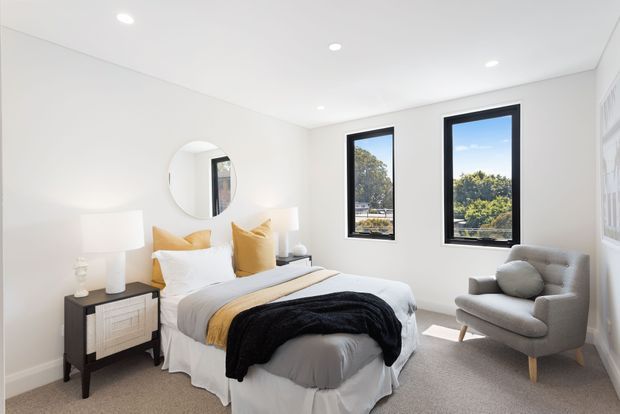 Bright, contemporary bedroom with a neatly styled bed, mustard and white cushions, layered grey and black throws, a dark bedside table, round wall mirror, upholstered armchair, recessed ceiling lights, and black-framed windows overlooking greenery.