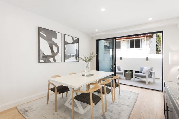Bright contemporary dining area opening directly onto a compact outdoor patio through large black-framed glazed doors. The room features a white rectangular dining table, light timber dining chairs with woven seats, pale timber flooring, a neutral rug, white walls, recessed ceiling lights, and abstract monochrome artwork. A small outdoor seating area and neighbouring building are visible beyond the glazing.