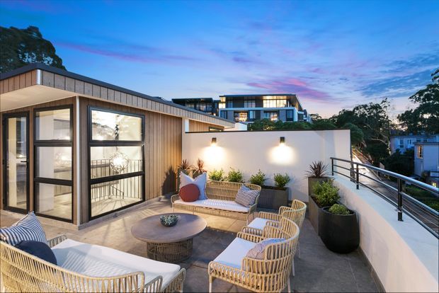 A contemporary rooftop terrace at dusk with a warm outdoor seating arrangement, including woven lounge chairs, a sofa, a round coffee table, large planters, and subtle wall lighting. The terrace has pale rendered walls, dark metal balustrades, timber-look vertical cladding, and views toward surrounding modern homes and trees.