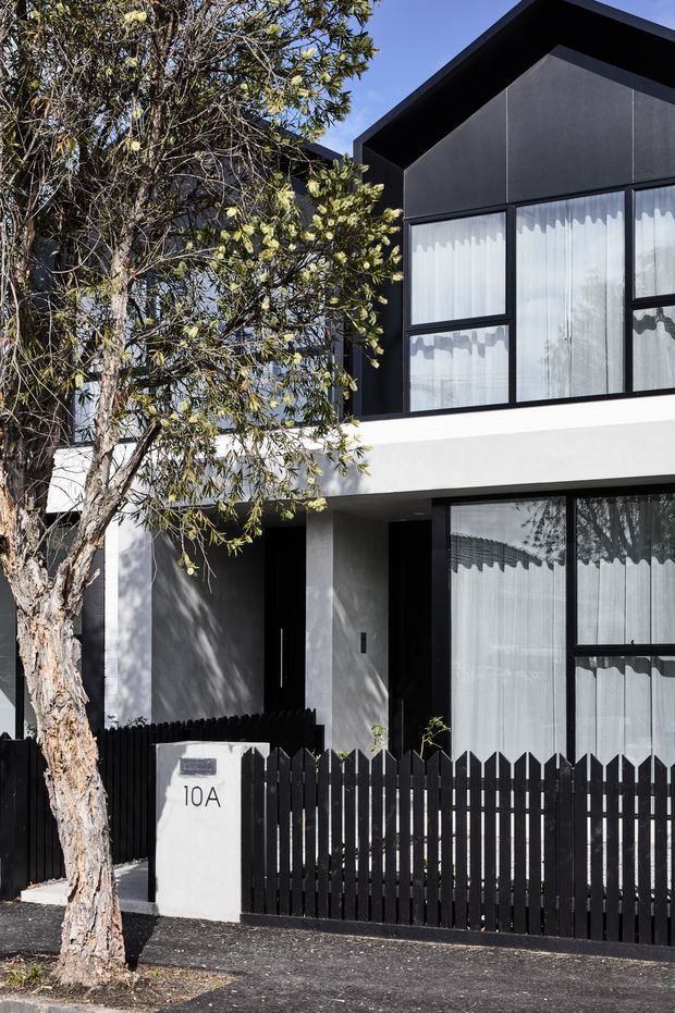 Street-facing view of a contemporary two-storey residence with a strong black-and-white facade. The upper level has a steep gabled roof and dark cladding surrounding large black-framed windows with sheer curtains. A white horizontal balcony or parapet band separates the floors, while the ground floor features dark recessed entry elements, expansive glazing, a black picket fence, and a white rendered mailbox marked 10A. A mature street tree partially obscures the facade.